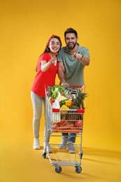 Young couple with shopping cart full of groceries on yellow background