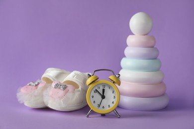 Alarm clock, toy pyramid and baby booties on violet background. Time to give birth