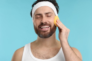 Photo of Man with headband washing his face using sponge on light blue background