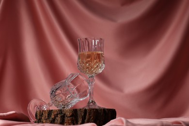 Glass of tasty alcoholic drink and empty one on wooden podium against color fabric