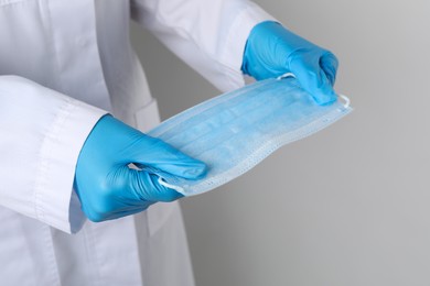 Doctor holding protective mask on light grey background, closeup