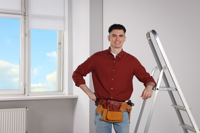 Young handyman with tool belt leaning on stepladder in room