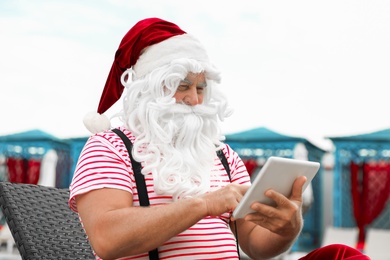 Photo of Authentic Santa Claus with tablet working on lounge chair at resort