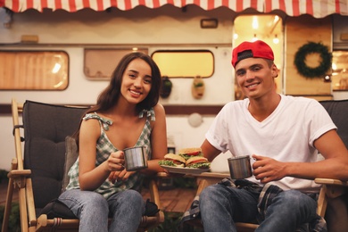 Photo of Happy couple with burgers and cups resting near trailer. Camping season