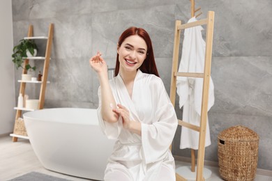 Photo of Beautiful young woman applying body cream onto elbow in bathroom