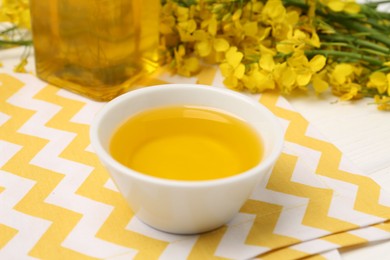 Photo of Rapeseed oil in bowl and beautiful yellow flowers on table, closeup