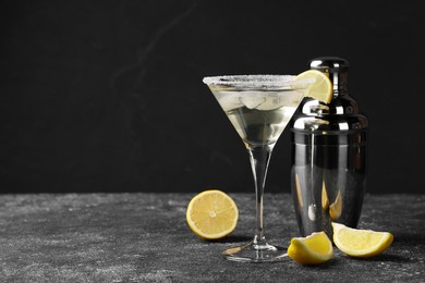 Photo of Martini glass of refreshing cocktail with lemon and ice cubes near shaker on black textured table. Space for text