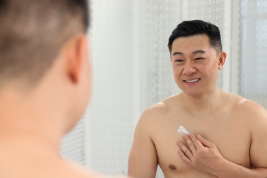 Photo of Man applying body cream onto his chest near mirror indoors