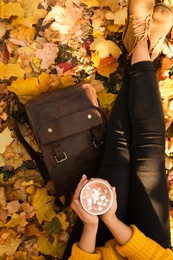 Photo of Woman holding cup of hot drink in park with fallen leaves, above view. Autumn season