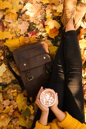 Woman holding cup of hot drink in park with fallen leaves, above view. Autumn season