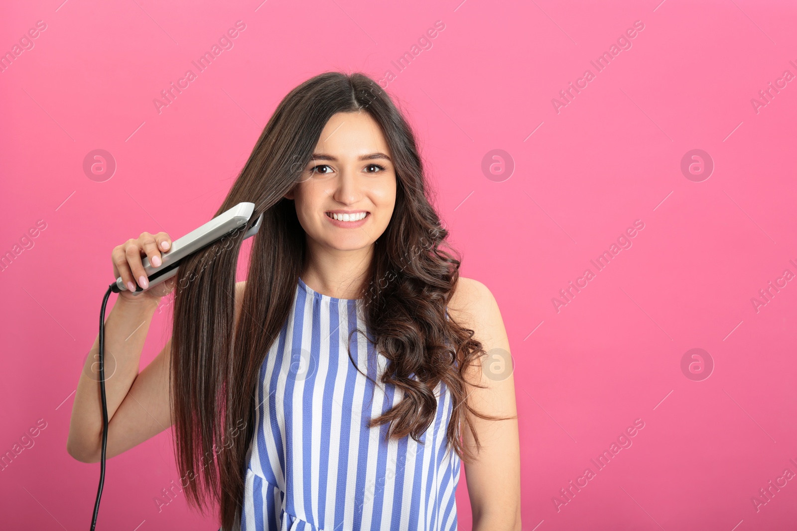 Photo of Young woman using hair iron on pink background, space for text