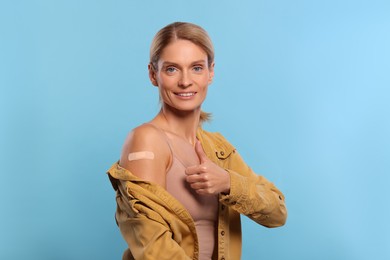Photo of Smiling woman with adhesive bandage on arm after vaccination showing thumb up on light blue background