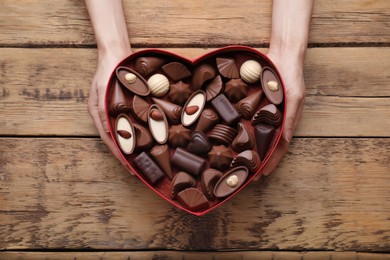 Woman holding heart shaped box with delicious chocolate candies at wooden table, top view