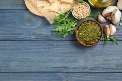 Photo of Bowl of tasty arugula pesto and ingredients on blue wooden table, flat lay. Space for text