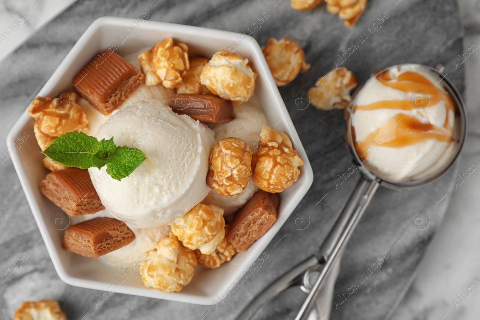 Photo of Bowl of delicious ice cream with caramel candies, popcorn and mint on grey marble board, flat lay