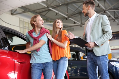 Photo of Salesman with clipboard consulting young couple in modern car dealership