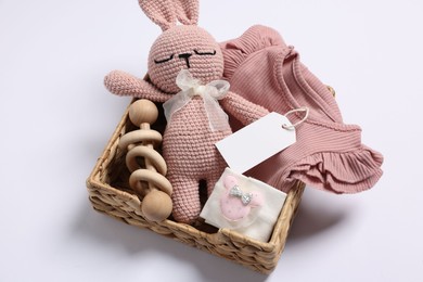 Different baby accessories, clothes and blank card in wicker box on white background