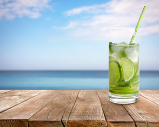 Tasty lemonade with ice cubes and lime on wooden table near sea, space for text