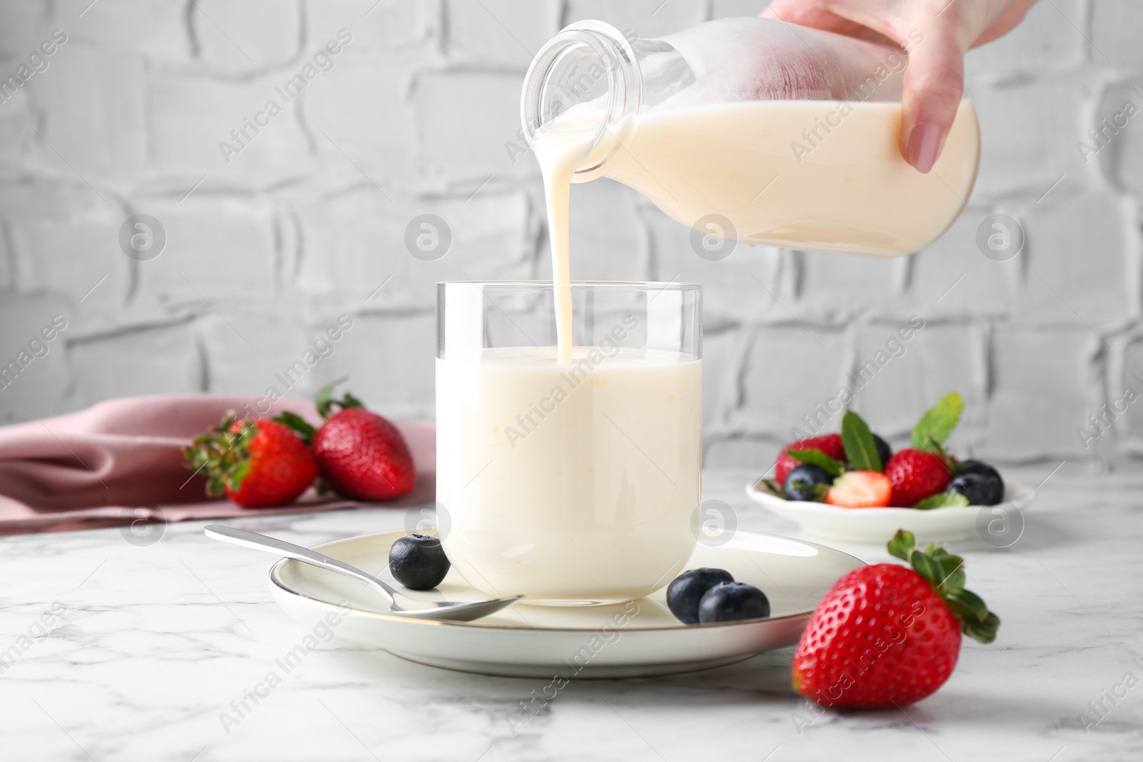 Photo of Woman pouring tasty yogurt into glass at white marble table, closeup