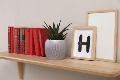 Photo of Wooden shelf with different books, houseplant and frames on light wall