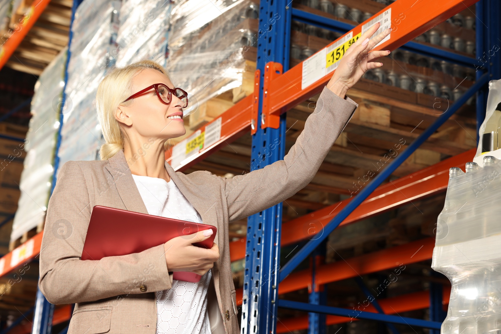 Photo of Happy manager using modern tablet and pointing at something in warehouse with lots of products, low angle view