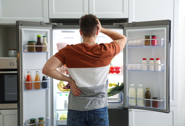 Photo of Young man near open refrigerator indoors, back view