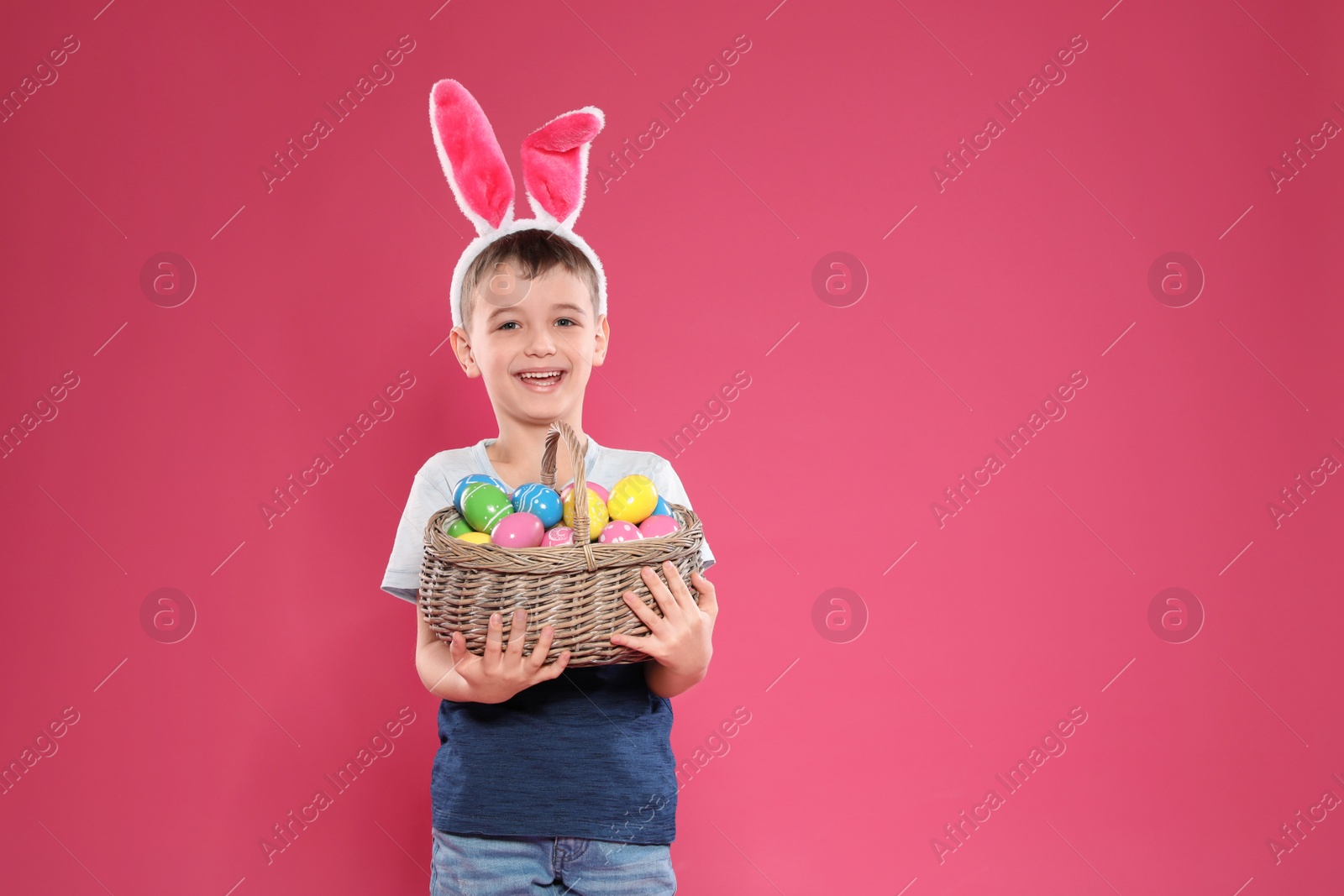 Photo of Little boy in bunny ears headband holding basket with Easter eggs on color background, space for text