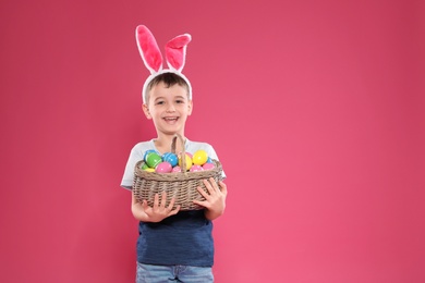 Little boy in bunny ears headband holding basket with Easter eggs on color background, space for text