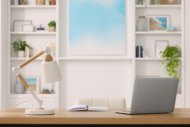 Photo of Interior design. Cosy workplace with laptop and lamp on wooden table near abstract picture between shelves