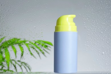 Bottle with moisturizing cream and palm leaves on light background, view through wet glass