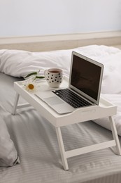 Photo of White tray table with laptop, cup of drink and daisy on bed indoors
