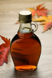 Photo of Glass bottle of tasty maple syrup and dry leaves on wooden table, closeup