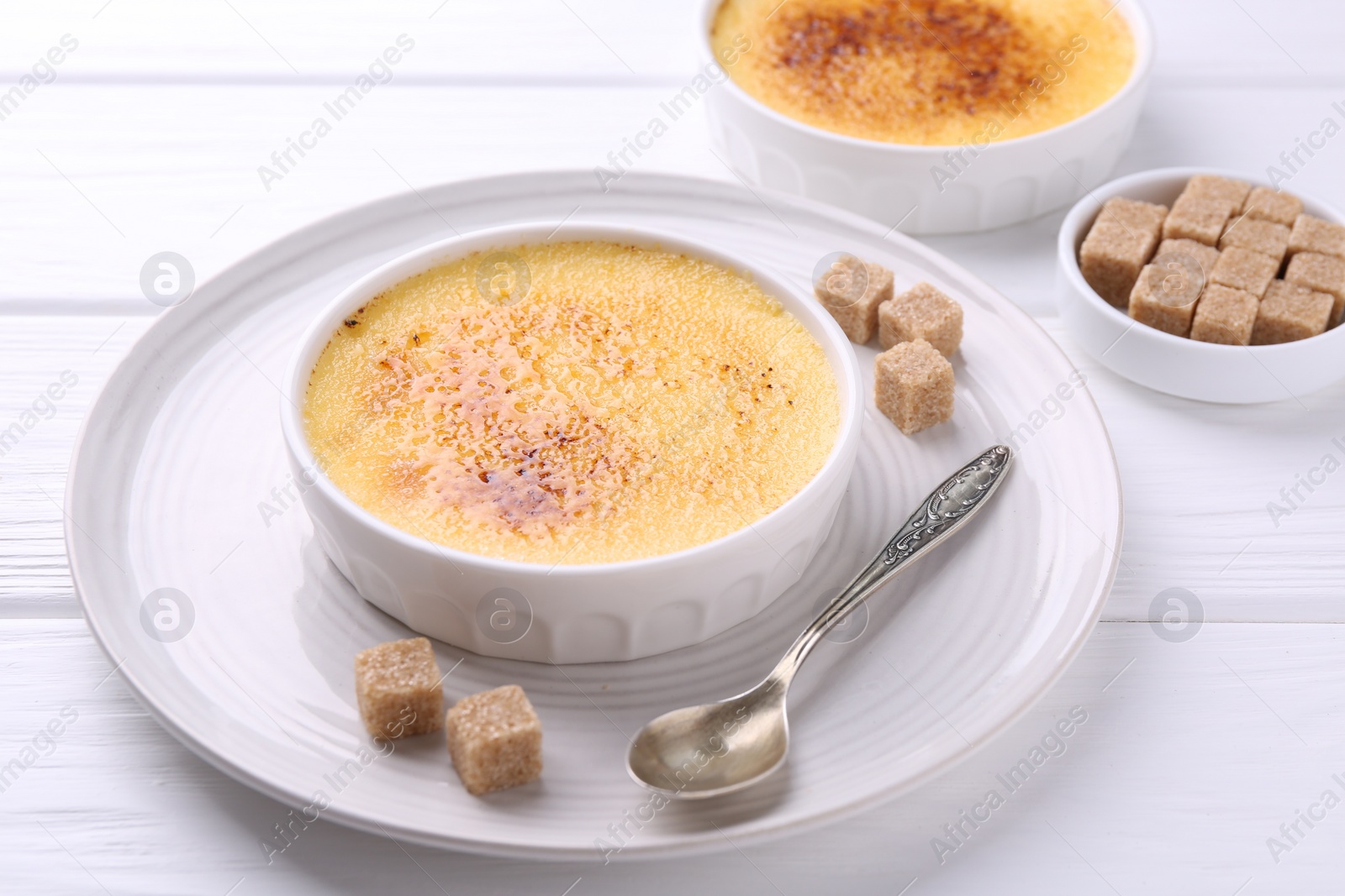 Photo of Delicious creme brulee in bowls, sugar cubes and spoon on white wooden table