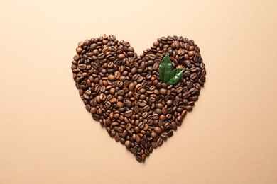 Photo of Heart shaped pile of coffee beans and fresh green leaves on light orange background, top view