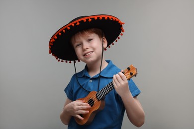 Photo of Cute boy in Mexican sombrero hat playing ukulele on grey background