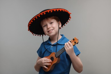 Cute boy in Mexican sombrero hat playing ukulele on grey background