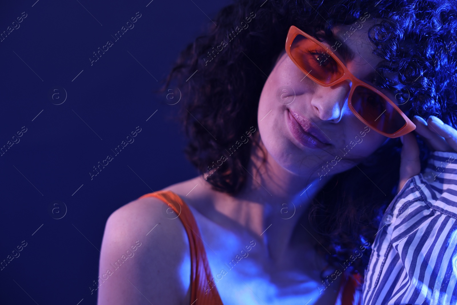 Photo of Beautiful young woman in sunglasses posing on color background in neon lights, closeup. Space for text