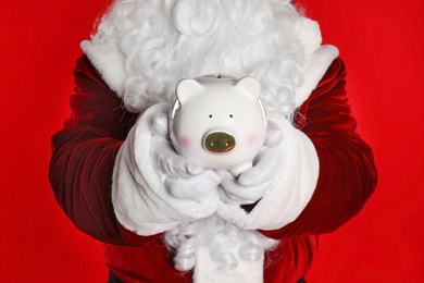 Photo of Santa Claus holding piggy bank on red background, closeup