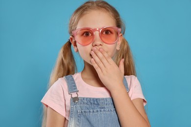 Photo of Girl in pink sunglasses covering mouth with hand on light blue background