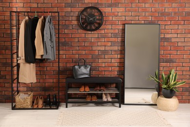 Stylish hallway with coat rack, mirror and shoe storage bench near brick wall. Interior design