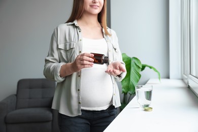 Pregnant woman taking pill from bottle near window at home, closeup