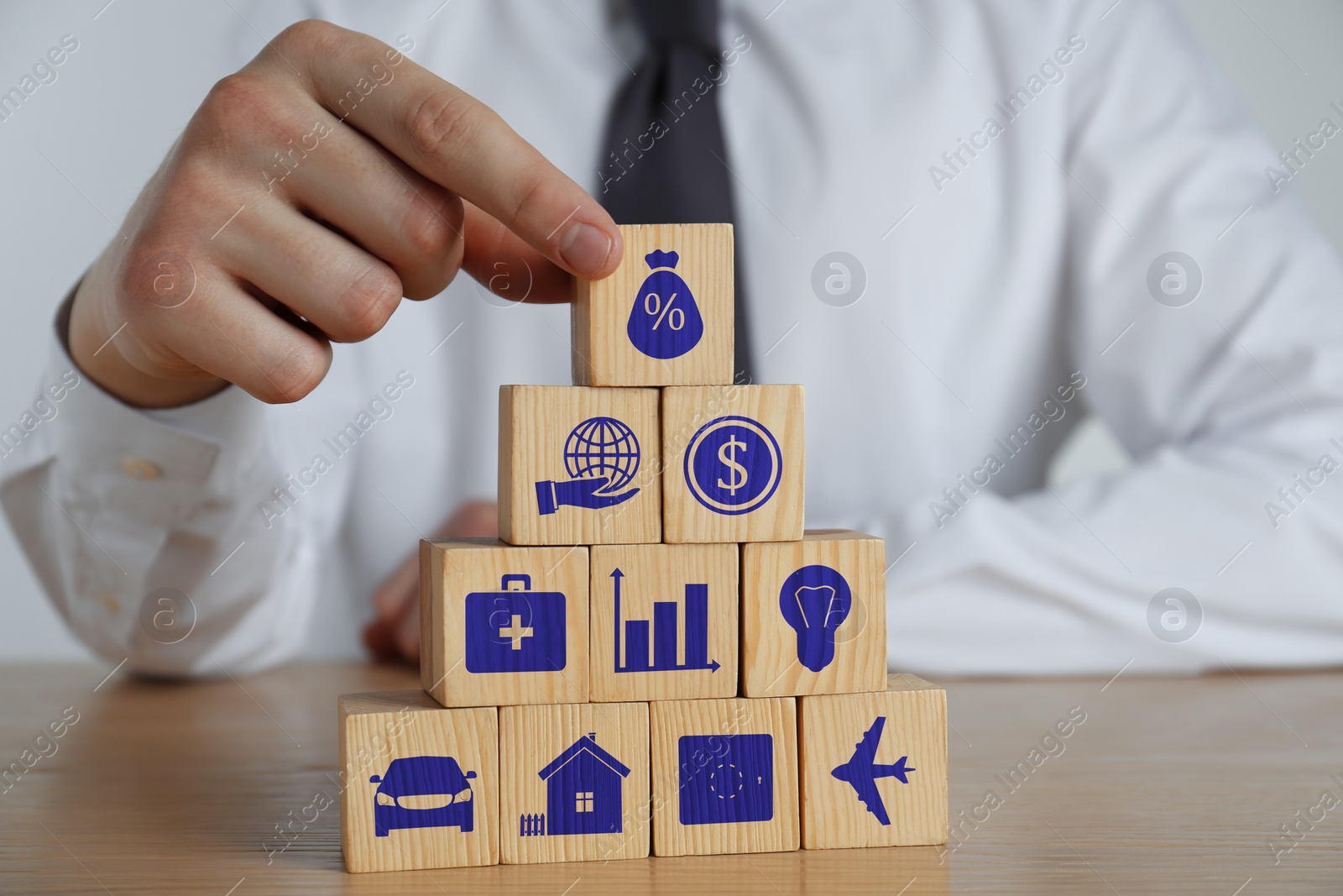 Image of Man building pyramid of cubes with different icons at wooden table, closeup. Insurance concept