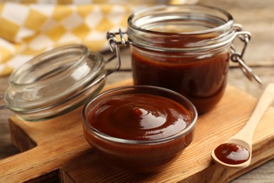 Photo of Tasty barbeque sauce in bowl, jar and spoon on wooden table, closeup