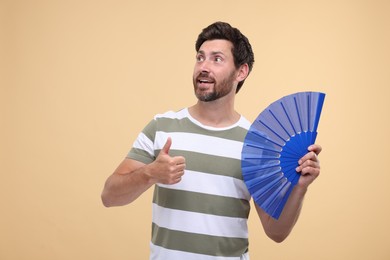 Happy man holding hand fan and showing thumb up on beige background