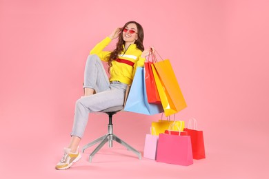 Happy woman in stylish sunglasses holding many colorful shopping bags on armchair against pink background