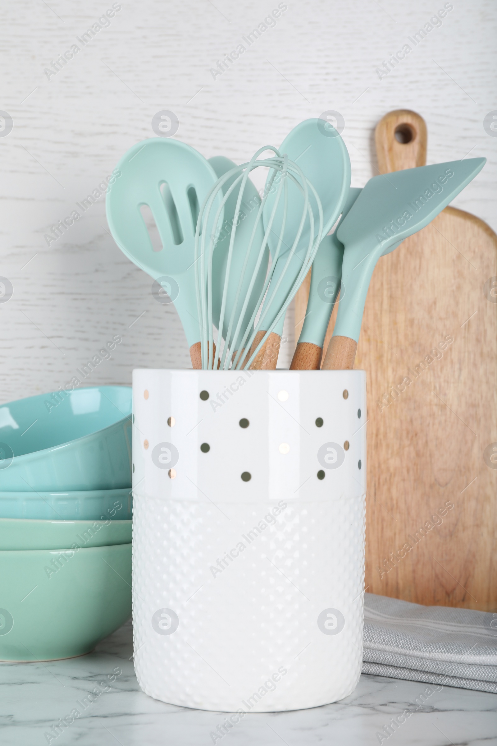 Photo of Holder with kitchen utensils, ceramic dishes and cutting board on white marble table