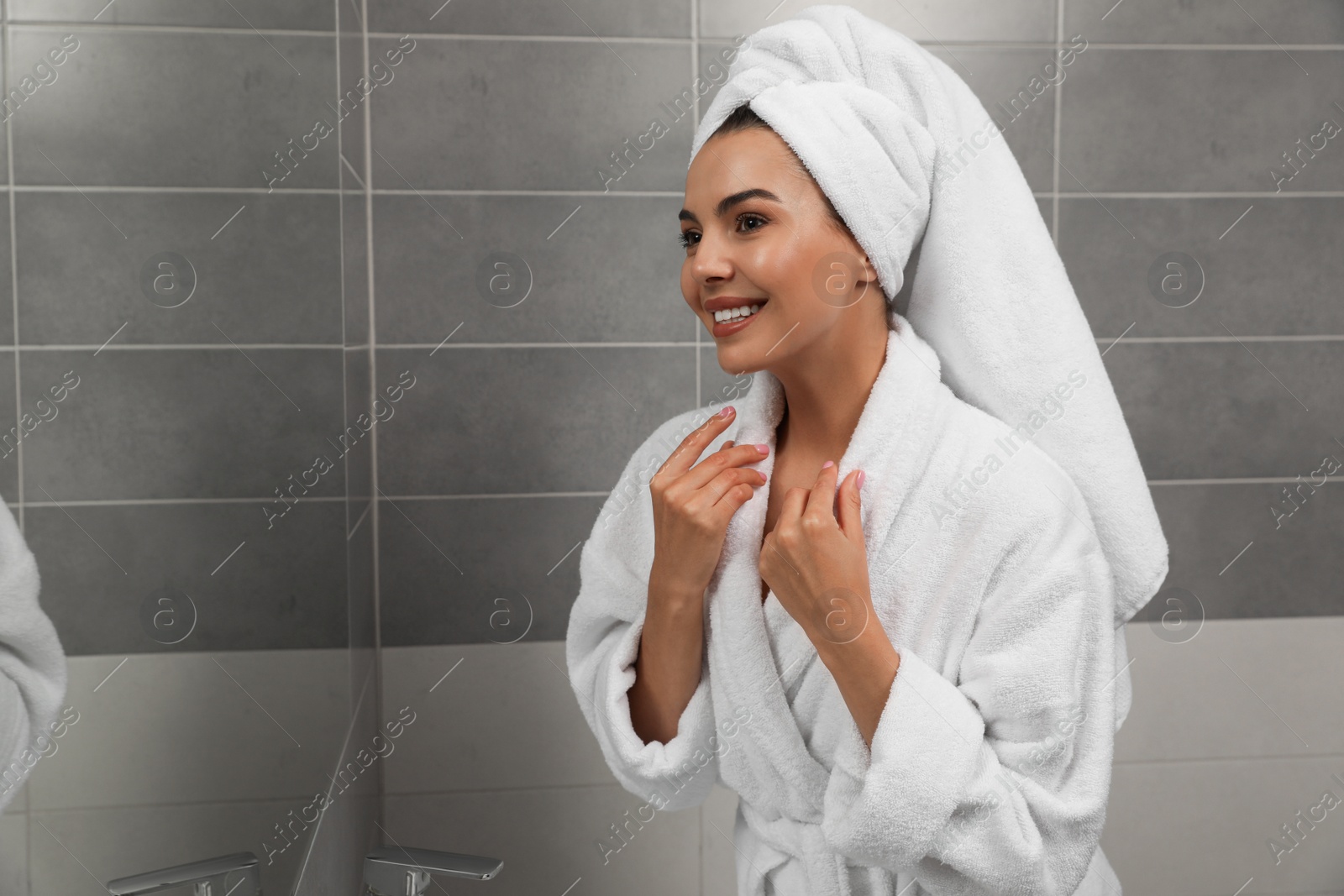 Photo of Beautiful young woman in bathrobe with towel on head near mirror at home