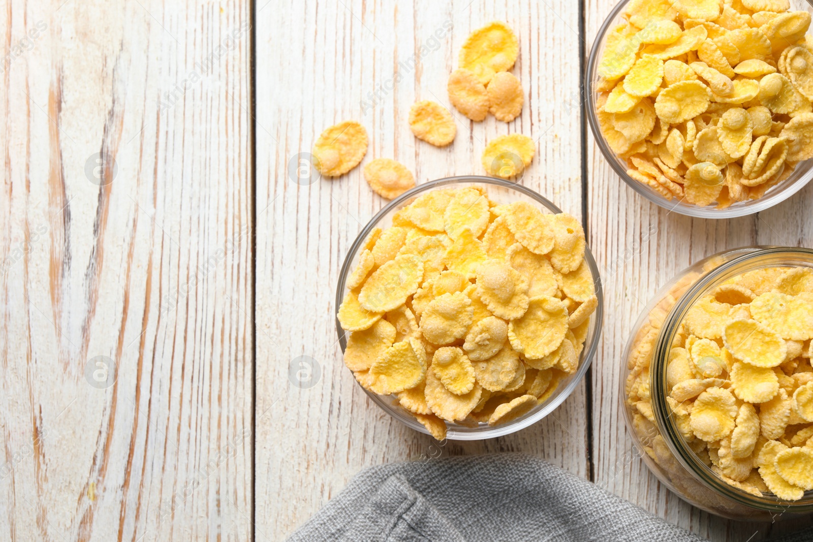 Photo of Glass bowls and jar of tasty crispy corn flakes on white wooden table, flat lay. Space for text