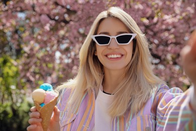 Photo of Happy young woman with icecream taking selfie near blossoming tree in park