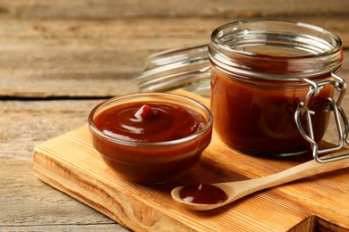 Photo of Tasty barbeque sauce in bowl, jar and spoon on wooden table, closeup